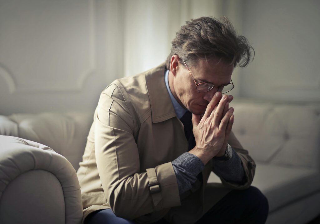 The image shows a middle-aged man sitting indoors on a light-colored couch, leaning forward with his elbows on his knees. He has his hands clasped together near his face, with his eyes closed and head bowed slightly, suggesting deep thought, worry, or prayer. He is wearing glasses, a beige trench coat over a blue shirt, and a wristwatch. The soft lighting and muted background create a calm yet somber atmosphere, conveying a mood of contemplation, stress, or emotional struggle.
