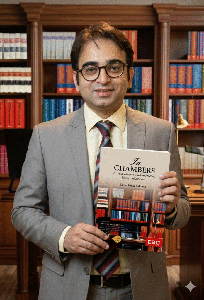 Famous lawyer talha abdul rahaman standing in his chembar wearing a gray suit and hold his new release book on his hand "in chembars"