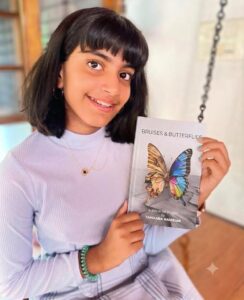 A young Indian girl smiling broadly while holding a copy of the book "Bruises & Butterflies" by Tamaara Nambiar. She is wearing a light purple ribbed turtleneck, a delicate necklace, and a green beaded bracelet. The background is a soft-focus indoor setting with wooden blinds and a chain. This image is perfect for a review of the book "Bruises & Butterflies" or a post about young authors.