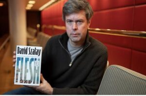 Author David Szalay holding his novel, Spring, against a red wall background.