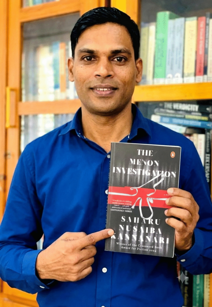 Man in blue shirt holding and pointing to the book "The Menon Investigation" by Saharu Nusaiba Kannanari in front of a bookshelf.